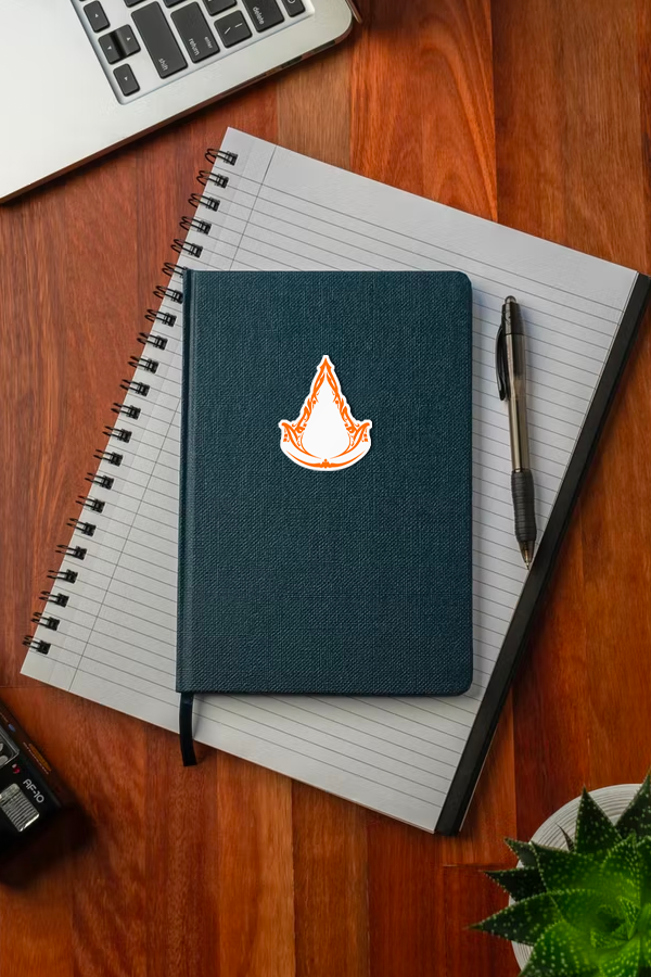 Image shows Assassin's Creed Mirage Decal stuck on the front cover of a journal. This Assassins Creed Mirage Vinyl Decal is perfect for sticking on your car, laptop, phone case, door, wall, or any other flat surface.